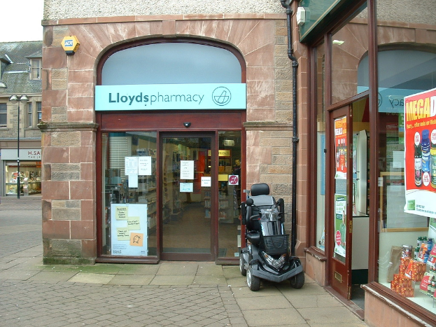 Eine Apothekenfiliale mit einem vor dem Gebäude geparkten Fahrzeug und einem Gebäude auf der linken Seite.