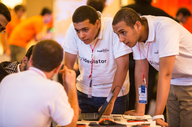 Mehrere Männer sitzen um einen Tisch mit einem Laptop, einer Maus auf einem Pad und einem angeschlossenen Kabel, alles leicht unscharf im Hintergrund.