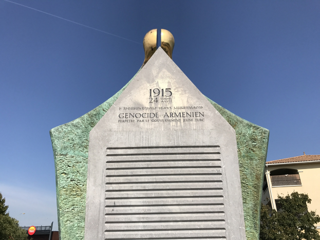 Ein steinernes Denkmal in einem Park mit einer Uhr darauf, umgeben von Bäumen und Gebäuden, mit einer Inschrift, die den 24. Jahrestag des Völkermords in Armenien erinnert, unter einem klaren Himmel.