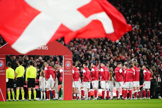 Gruppe von Menschen auf einem Fußballfeld mit einer roten und weißen Flagge im Vordergrund, einem Bogen mit dem Text "Bayern München vs. Bayern München Wetten & Vorschau" im Hintergrund und einer großen Menschenmenge im Stadion.