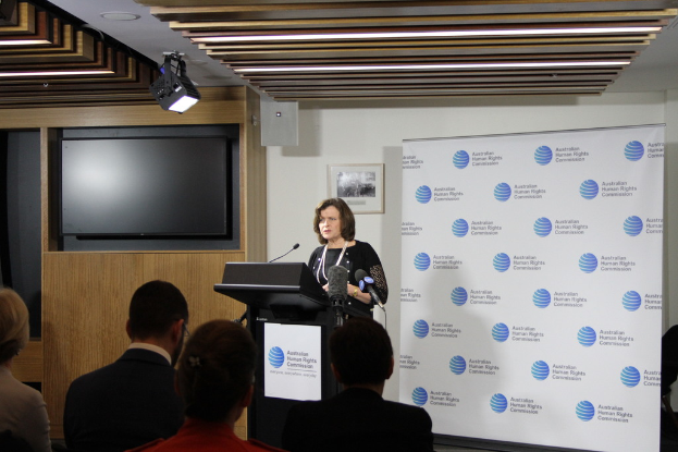 Eine Frau steht auf einer Bühne und spricht in ein Mikrofon, vor ihr sitzen Zuschauer, an der Wand hängt ein Banner, ein Fernseher steht an der Holzwand, und an der Decke sind Lampen.