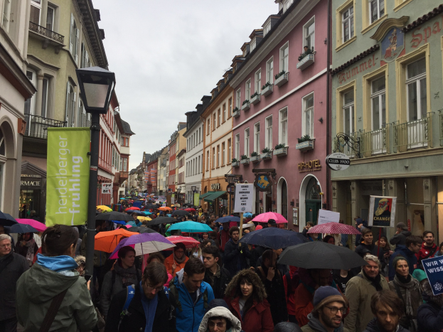 Große Gruppe von Menschen, die mit Schirmen und Plakaten auf einer Straße in Deutschland marschieren.