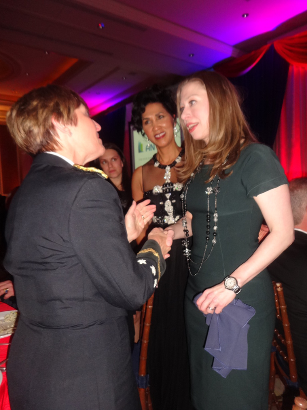 Vier Frauen im Vordergrund, die miteinander sprechen, mit einem Stuhl rechts, einem Vorhang oben rechts, Lampen oben und einem roten Objekt links.