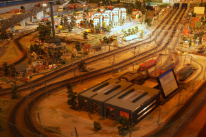 Eine detaillierte Miniatur-Modellbahnausstellung in einem Museum, die Gebäude, Bäume, Eisenbahnschienen, Fahrzeuge, Masten, Lichter und andere Gegenstände umfasst, die eine belebte Stadtlandschaft nachahmen.