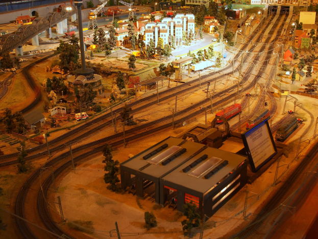 Eine detaillierte Miniatur-Modellbahnausstellung in einem Museum, die Gebäude, Bäume, Eisenbahnschienen, Fahrzeuge, Masten, Lichter und andere Gegenstände umfasst, die eine belebte Stadtlandschaft nachahmen.