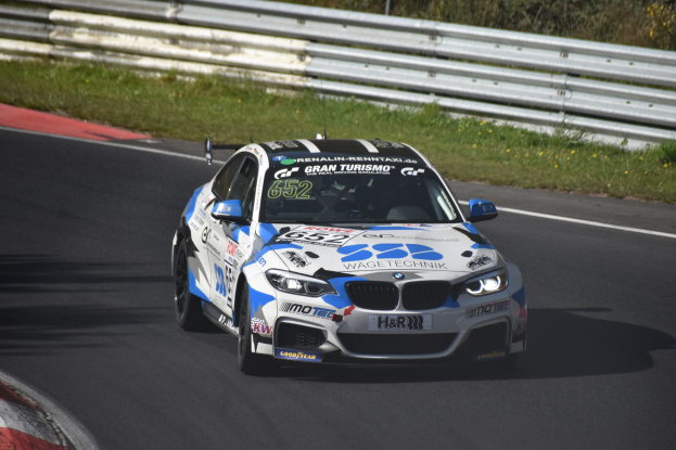 Ein BMW M4 DTM-Rennwagen fährt auf einer Rennstrecke, umgeben von Gras, einem Zaun und Pflanzen, mit Text und Zahlen auf seiner Karosserie und einem sichtbaren Rad.