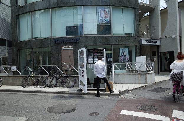 Eine Frau fährt auf einer Straße mit einem Fahrrad, während ein Mann in der Nähe einer Bude und mehrere Fahrräder in der Nähe eines Ladens stehen.