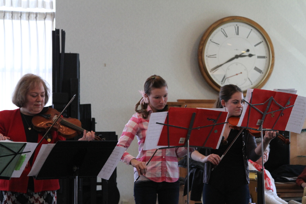 Drei Frauen stehen auf der Bühne und spielen Geigen an Musikständern, während eine Gruppe sitzender Frauen im Vordergrund zuhört; eine Uhr hängt an der weißen Wand im Hintergrund, mit einem weißen Vorhang in der linken Ecke.