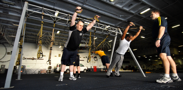 Eine Gruppe von Männern, die Klimmzüge in einem Fitnessstudio machen, wobei ein Mann in der Mitte Schuhe trägt und von Metallstangen, Rahmen, einer Wand, einem Dach mit Deckenleuchten und verschiedenen Gegenständen auf der Oberfläche umgeben ist.