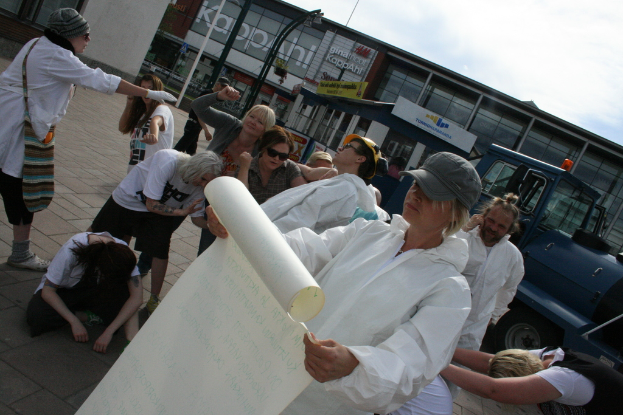Eine Gruppe von Menschen, einige in Kostümen und Mützen, versammelt sich in der Mitte, wobei einige zu kämpfen scheinen, während eine Person ein Papier in den Vordergrund hält. Im Hintergrund sind Gebäude, Pflanzen, Pfosten und Schilder mit Text, ein Fahrzeug auf der rechten Seite, ein Weg am Boden und der Himmel oben zu sehen.