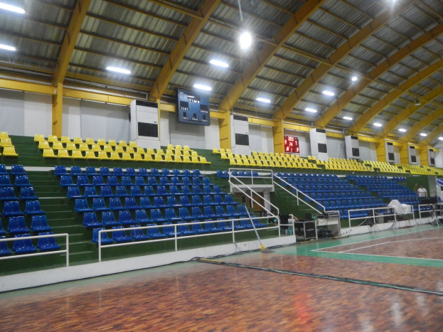 Großer Hallensportplatz mit Holzboden, blauen und gelben Sitzplätzen, Geländern, Treppen mit Geländern, wandmontierten Bildschirmen, Deckenleuchten und wenigen Menschen.
