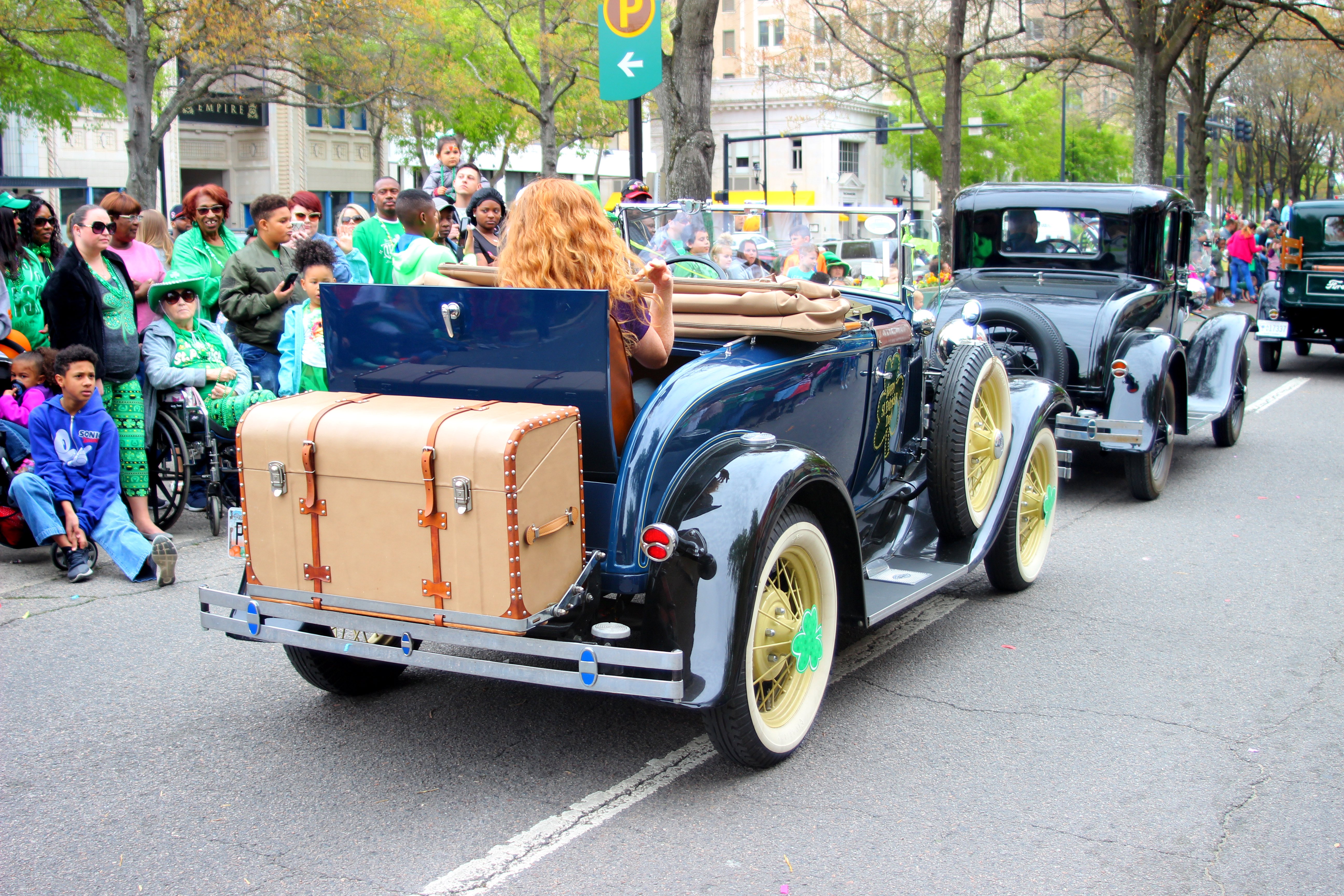 Eine Gruppe von Menschen im R├╝cken eines Oldtimers mit einer Frau am Steuer, Koffern und einer Person im Rollstuhl auf der linken Seite, mit B├Ąumen, Geb├Ąuden und Schildern im Hintergrund.
