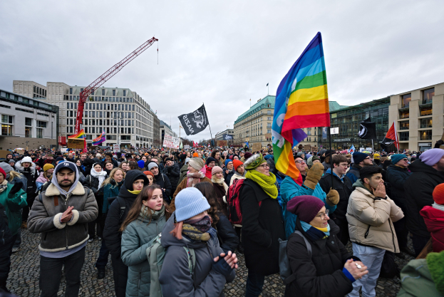 Eine große Gruppe von Menschen steht vor einem Gebäude und hält Fahnen und Schilder mit der Aufschrift "Lgbtq+ Rechte Demonstration in Berlin", einige tragen Mützen und Taschen, vor einem Hintergrund aus Gebäuden, einem Kran und einem bewölkten Himmel.