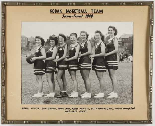 Schwarzes und weißes Foto einer Gruppe von Frauen, die lächeln und einen Basketball halten, mit der Aufschrift "Kodak Basketball Team Semi-Final 1949" oben und unten, umgeben von Menschen, Bäumen, Gebäuden und dem Himmel.