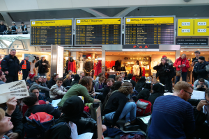 Eine große Gruppe von Menschen versammelt sich in einem Flughafen, einige sitzen mit Taschen und Papieren, andere stehen, mit Texttafeln, Schaufensterpuppen in Kleidern und Deckenleuchten im Hintergrund.