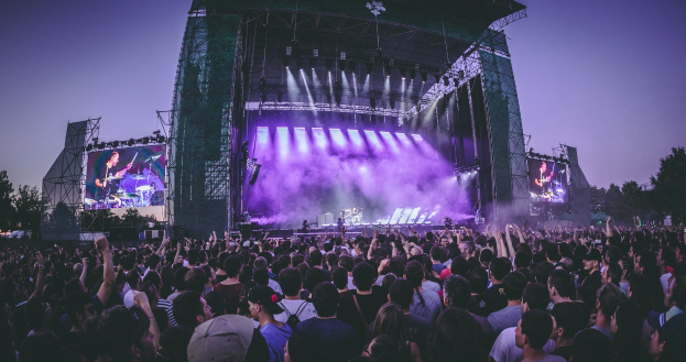 Eine große Menge bei einem Musikfestival schaut auf einer hell erleuchteten Bühne mit Instrumenten und Bildschirmen, die vor einem Gebäude, Bäumen und einem blauen Himmel steht.
