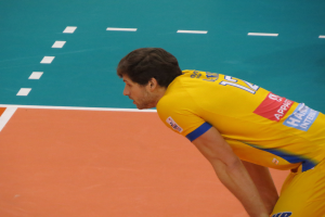 Ein Mann in einem gelben "Volleyball"-T-Shirt steht auf einem Volleyballfeld und ist bereit, den Ball mit Selbstvertrauen und Entschlossenheit zu servieren.