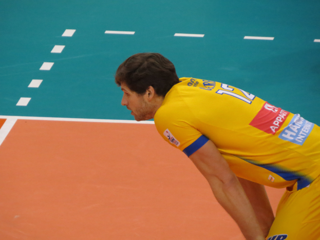 Ein Mann in einem gelben "Volleyball"-T-Shirt steht auf einem Volleyballfeld und ist bereit, den Ball mit Selbstvertrauen und Entschlossenheit zu servieren.