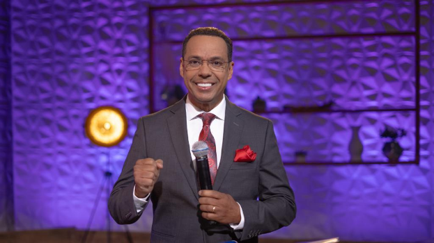 Stephen Colbert in Anzug, Krawatte und Brille auf einer Bühne mit Mikrofon, Wand, Ständer, Licht und diversen Gegenständen im Hintergrund.