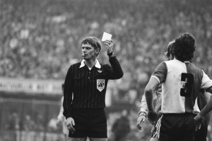 Schwarß-weißes Bild eines Schiedsrichters mit roter Karte vor einer Menge in Fußballkleidung, mit einem Zaun im Hintergrund.