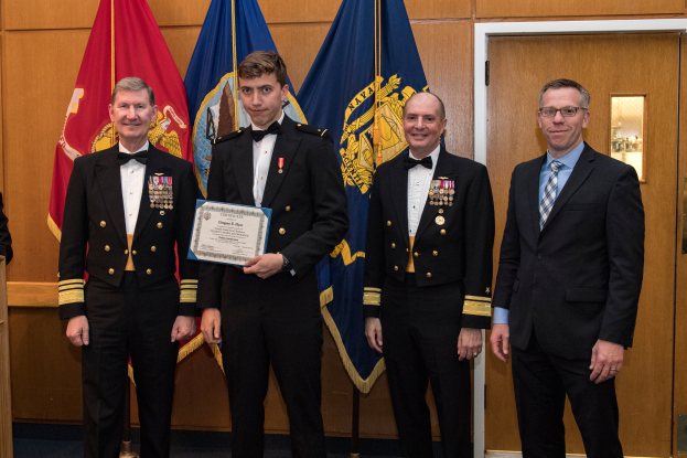 Eine Gruppe von Männern in Militäruniformen steht zusammen, einer hält ein Zertifikat, mit einem Podium und Mikrofon links, Flaggen und einer hölzernen Wand mit Tür im Hintergrund.
