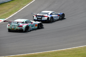Zwei Sportwagen auf einer Piste mit Gras an beiden Seiten und einem Geländer im Hintergrund