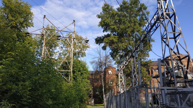 Gro├če Strommast mit mehreren Dr├Ąhten, die durch einen bewaldeten Bereich verlaufen, umgeben von B├Ąumen und einem Zaun, mit im Hintergrund sichtbaren Geb├Ąuden unter einem bew├Âlktem Himmel.