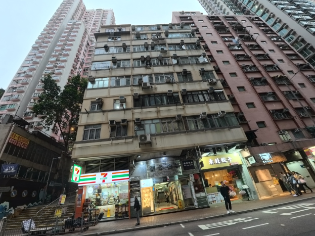 Eine belebte Straßen Szenerie mit hohen Gebäuden, Menschen auf dem Gehweg, Geschäften, Namensschildern, Strommasten, Stromkabeln, Bäumen und einem klaren blauen Himmel, mit einem Wohngebäude im Vordergrund.