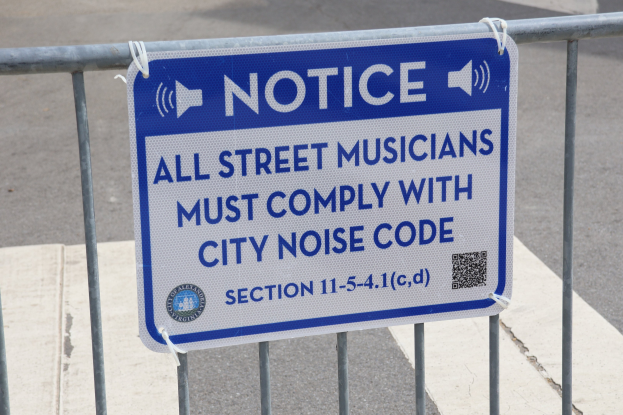 Anschlagtafel an einem Zaun mit einem Schild, auf dem "Anschlag: Alle Straßenmusikanten müssen die Stadtlärmordnung einhalten" steht, und einer Straße im Hintergrund.