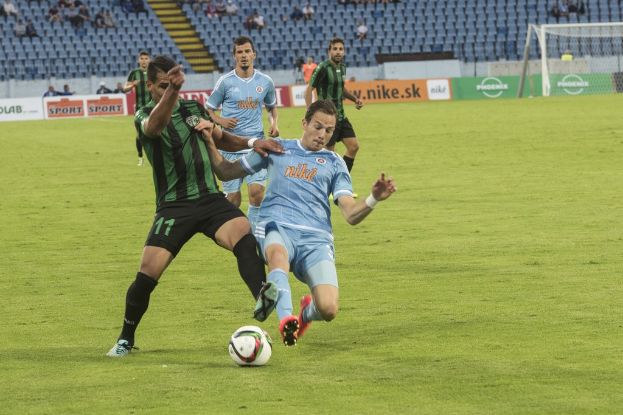 Outdoor game with players, a ball on grass, a net, hoardings, audience, stadium, and chairs in the background.