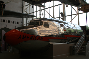 Ein Flugzeug von American Airlines in einem Museum mit Menschen drumherum, Treppe auf der rechten Seite und einem Gebäude, Bäumen und klarem blauen Himmel im Hintergrund.