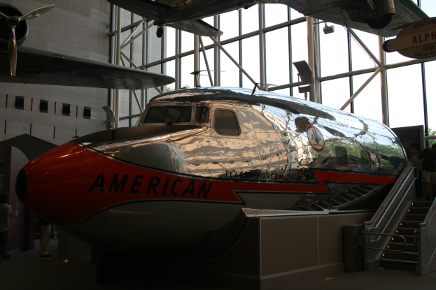 Ein Flugzeug von American Airlines in einem Museum mit Menschen drumherum, Treppe auf der rechten Seite und einem Gebäude, Bäumen und klarem blauen Himmel im Hintergrund.