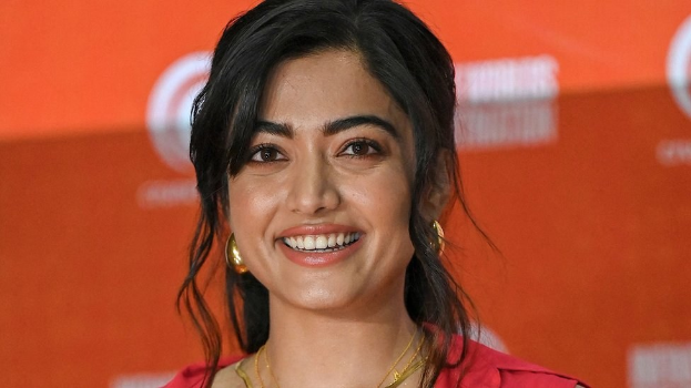 Eine Frau in einem roten Kleid mit einer eleganten Hochsteckfrisur und dezentem Make-up lächelt in die Kamera, trägt schwarze High Heels und hat ein leuchtend gelbes Banner mit der Aufschrift 'Die besten Schönheits Looks von Sonam Kapoor auf dem Cannes Film Festival' im Hintergrund.