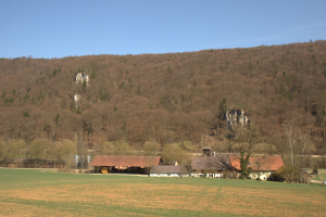 Bauernhof in einer grünen Landschaft mit Gebäuden, Bäumen und Bergen im Hintergrund unter einem blauen Himmel.