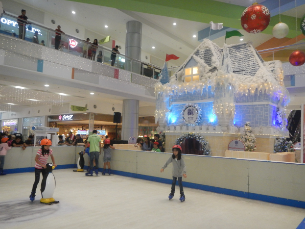 Eine Gruppe von Menschen, die auf einer Eisbahn in einem Einkaufszentrum Schlittschuh laufen, umgeben von festlichen Dekorationen, einschließlich eines hausähnlichen Gebäudes mit Lichtern und Weihnachtsornamenten.