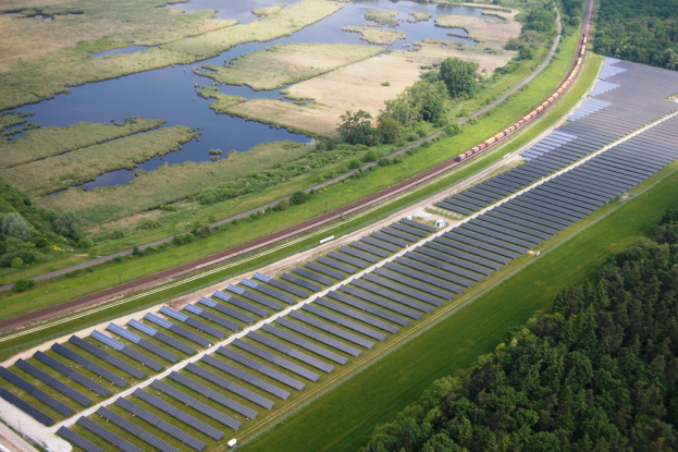 Luftaufnahme einer großen Solarstromanlage in einem Feld mit Bäumen, Gras, Pflanzen und Wasser, mit Solarpanelen und einem Zug auf einem Bahngleis.