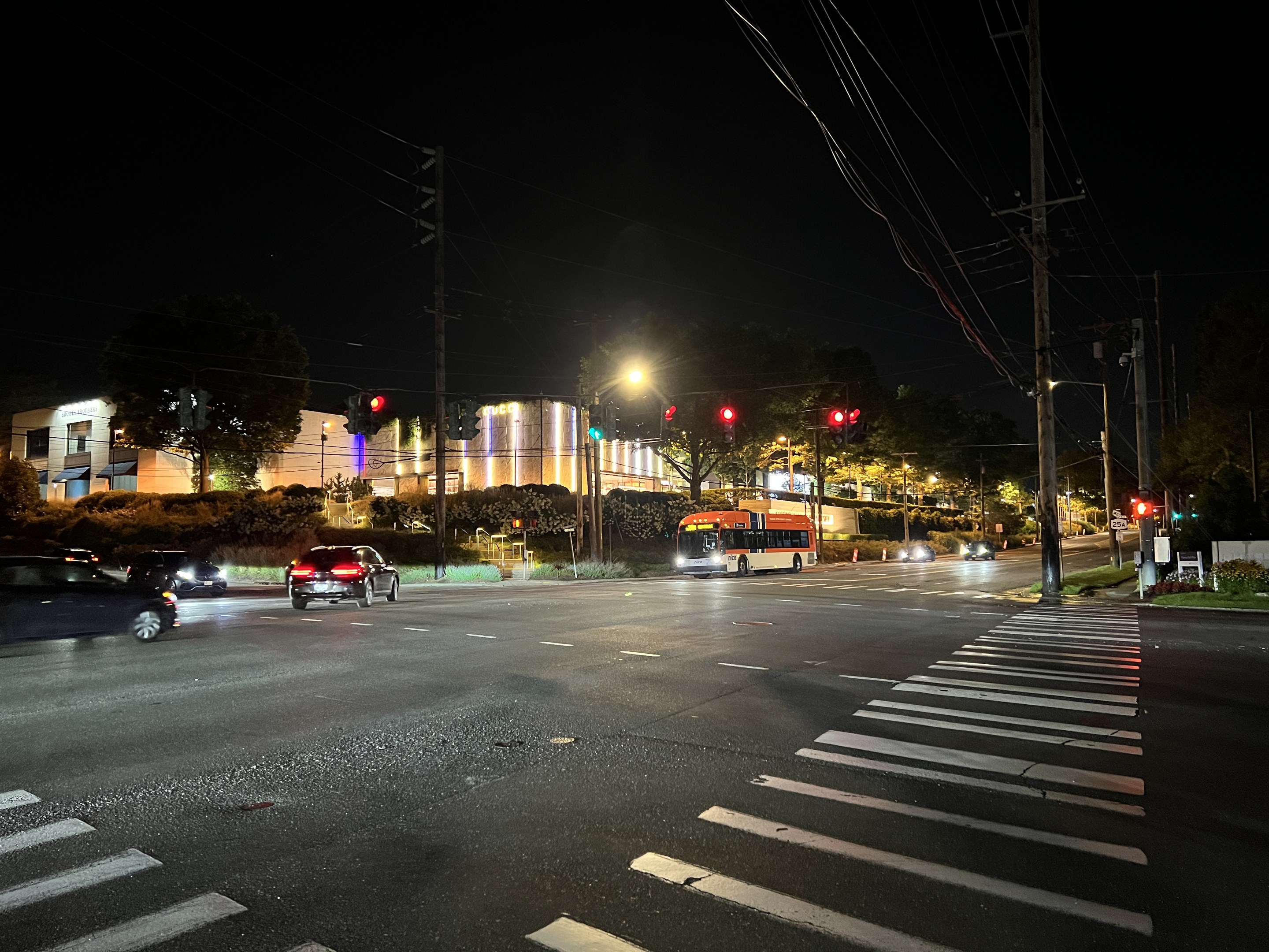 Eine nächtliche Stadtstraße mit fahrenden Autos und einem Bus, beleuchtet von Straßenlaternen und Ampeln, gesäumt von Bäumen, Gebäuden und Strommasten mit Überlandleitungen.