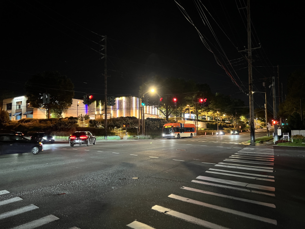 Eine nächtliche Stadtstraße mit fahrenden Autos und einem Bus, beleuchtet von Straßenlaternen und Ampeln, gesäumt von Bäumen, Gebäuden und Strommasten mit Überlandleitungen.