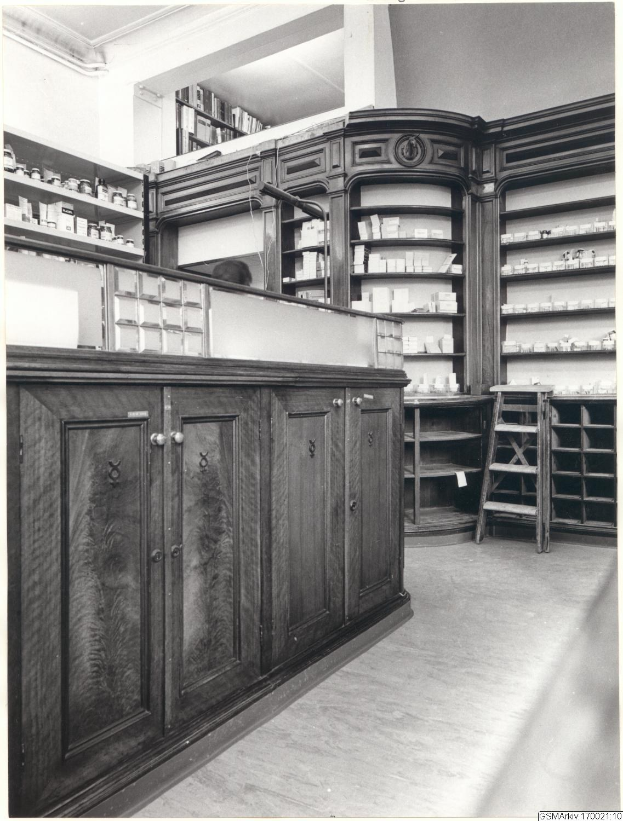 Schwarz-weiß-Foto eines alten Apothekeninneren mit einer Theke, Schränken, einer Leiter und Regalen, die mit verschiedenen Gegenständen gefüllt sind, mit Text oben.
