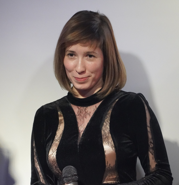 Frau in einem schwarzen Kleid, die ein Mikrofon hält, vor einer Wand im Hintergrund.