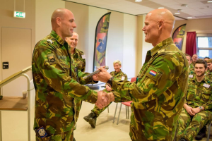 Zwei Männer in deutscher Militäruniform schütteln sich die Hände vor einer Gruppe von Menschen, die auf Stühlen sitzen, mit einem Tisch, Bannern und anderen Raumdetails im Hintergrund.
