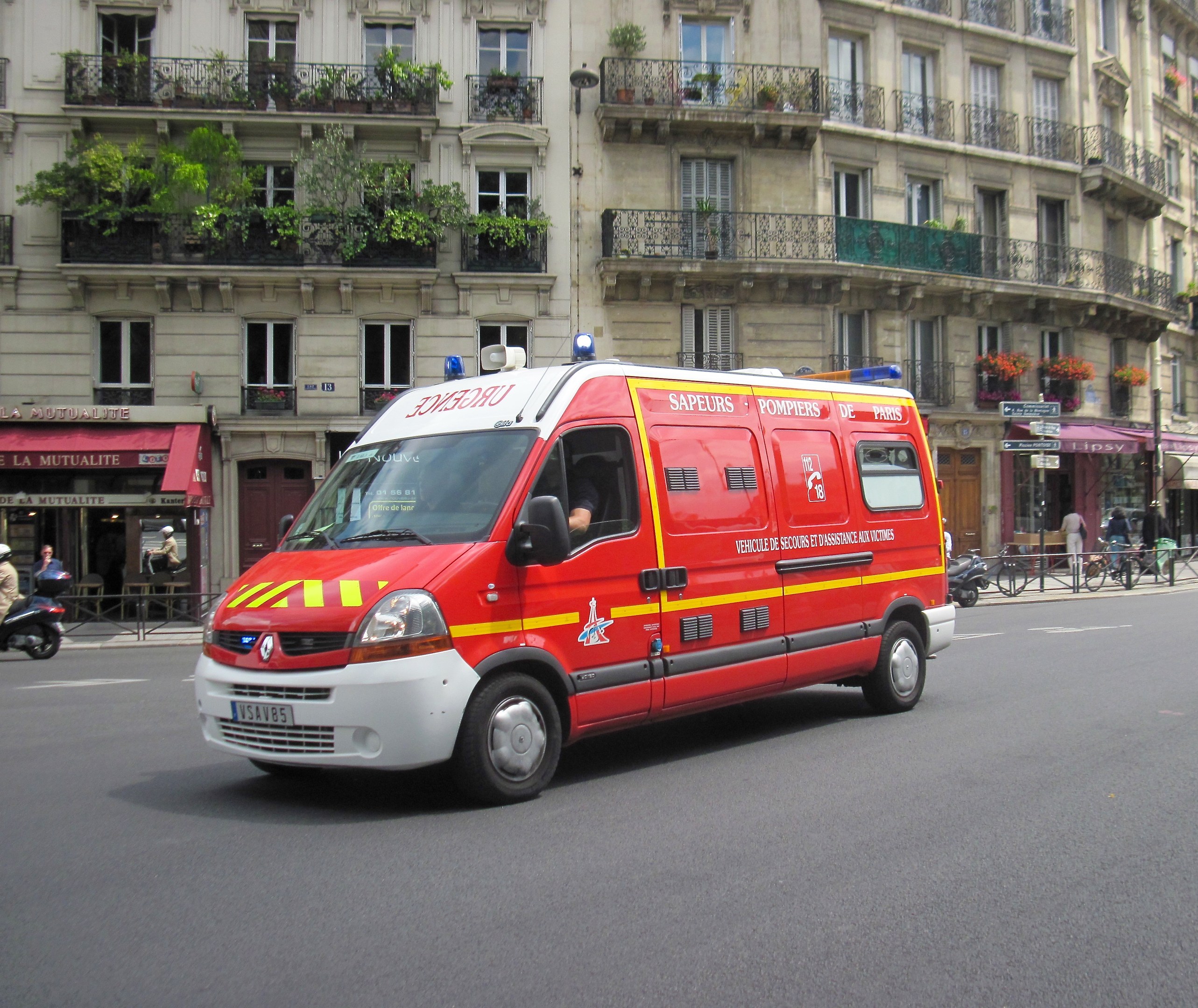 Ambulanz auf der Straße neben einem Gebäude mit Fenstern, Geländern, Pflanzen und anderen Gegenständen geparkt, mit ein paar Menschen und Fahrrädern im Hintergrund.