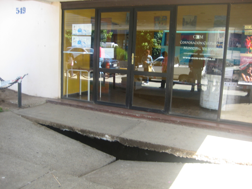 Eine gebrochene Oberfläche im Vordergrund mit einer Glastür, Wand und Stange im Hintergrund, die sich bewegende Fahrzeuge und einige stehende Personen reflektiert.