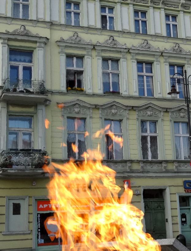 Ein großes Feuer brennt vor einem Gebäude mit Fenstern, Türen, einem Laternenpfahl und einer Tafel mit Schrift darauf.