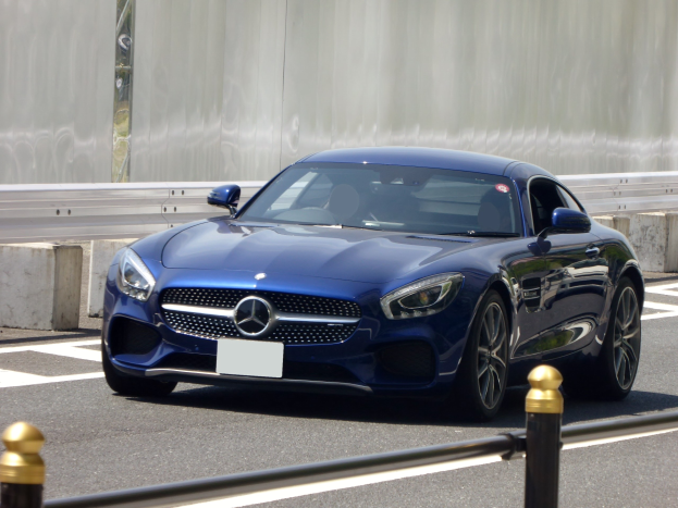 Blauer Mercedes-Benz AMG GT Coupé fährt auf einer Straße mit einer Wand im Hintergrund, mit einem eleganten aerodynamischen Design, eckigen Scheinwerfern, einem großen Kühlergrill, glänzenden großen Rädern und einem luxuriösen Lederinnenraum mit einem modernen Armaturenbrett.