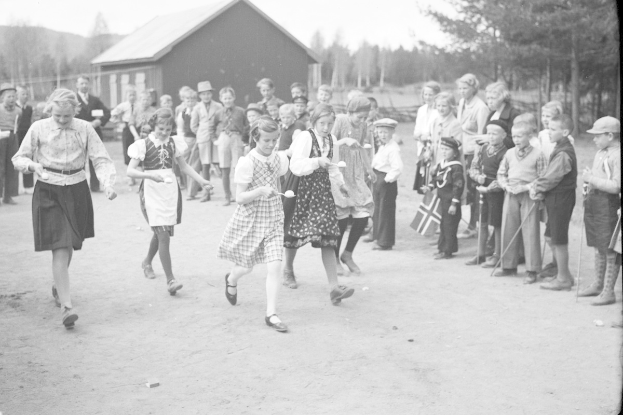 Schwarzes und weißes Bild von Kindern, die vor einer Menge mit Fahnen Fußball spielen, mit Bäumen und einem Haus im Hintergrund.