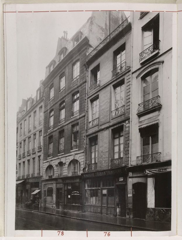 Ein Schwarz-Weiß-Foto einer Straße in einer Stadt mit Gebäuden, die Fenster, Geländer und Namensschilder aufweisen, mit einer Straße unten.