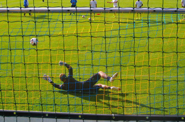 Ein Fußballspieler macht eine Parade vor einem Tor, mit einem Netz im Vordergrund und einigen Menschen im Hintergrund sowie mehreren Bällen auf dem Boden verstreut.