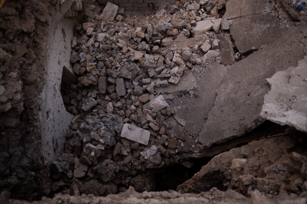 Zusammengebrochenes Gebäude mit einem großen Loch im Boden, umgeben von Trömmern und einer sichtbaren Leitung auf der linken Seite, was auf Erdbebenschäden hinweist.