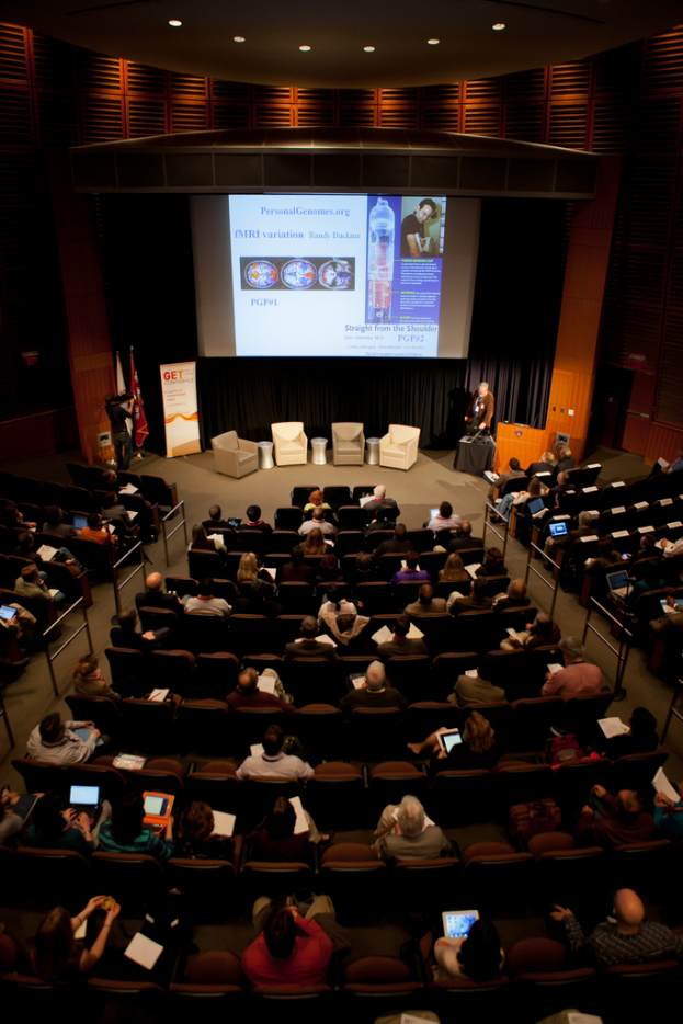 Ein mit Sitzenden beleuchteter Saal mit Fahnen, Rednerpult und Werbetafel